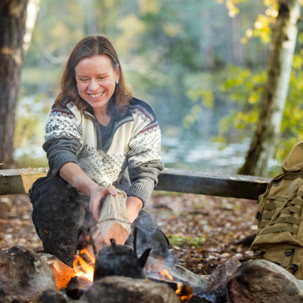 Man and woman making coffee in big kettle on campfire in forest on shore of lake, making a fire, grilling. Happy couple exploring Finland. Scandinavian landscape.